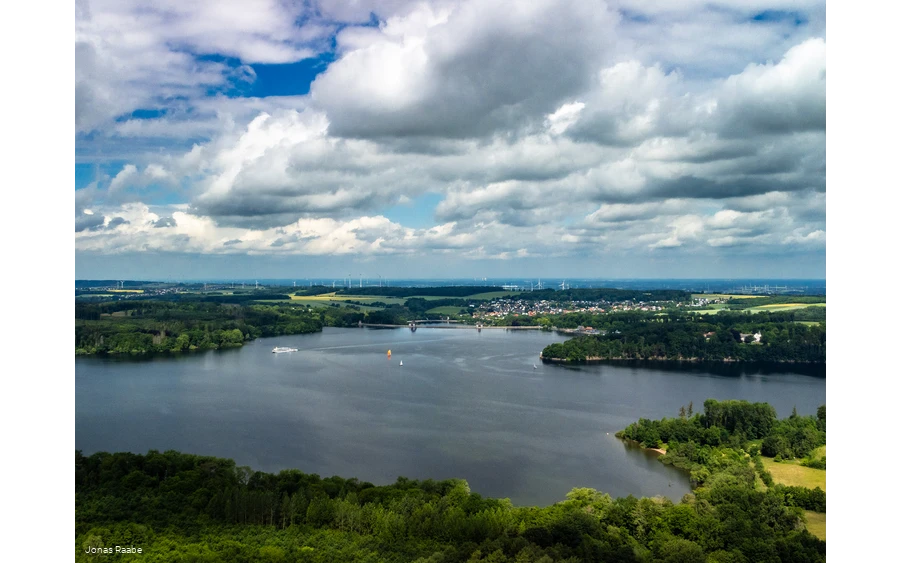 Luftaufnahme des Möhnesees mit Segelbooten und umliegender Landschaft unter Wolkenhimmel.