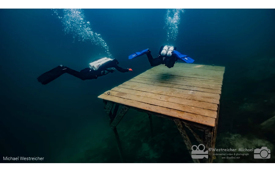 Zwei tauchende Personen erkunden einen versunkenen Steg unter Wasser.