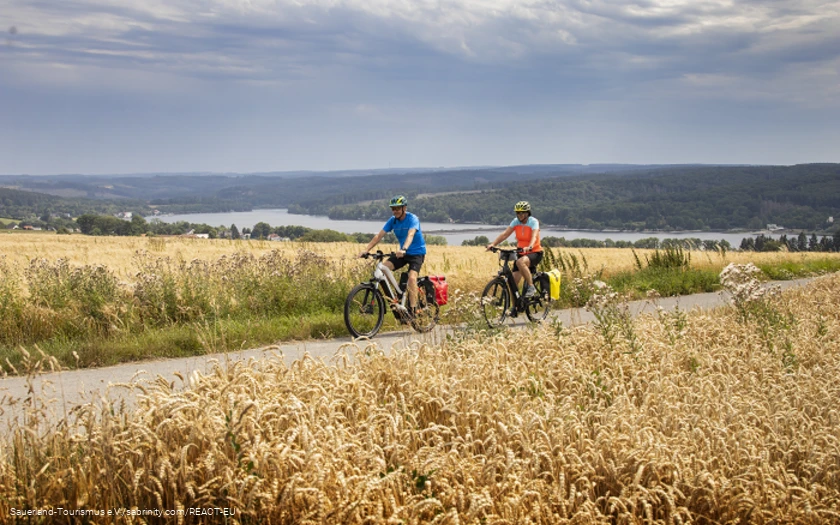 Zwei Radfahrer auf einem Weg durch Getreidefelder mit weiter Landschaft und dem Möhnesee im Hintergrund.
