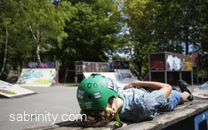 Junge Person mit grünem Helm liegt auf einer Bank in einem Skatepark.