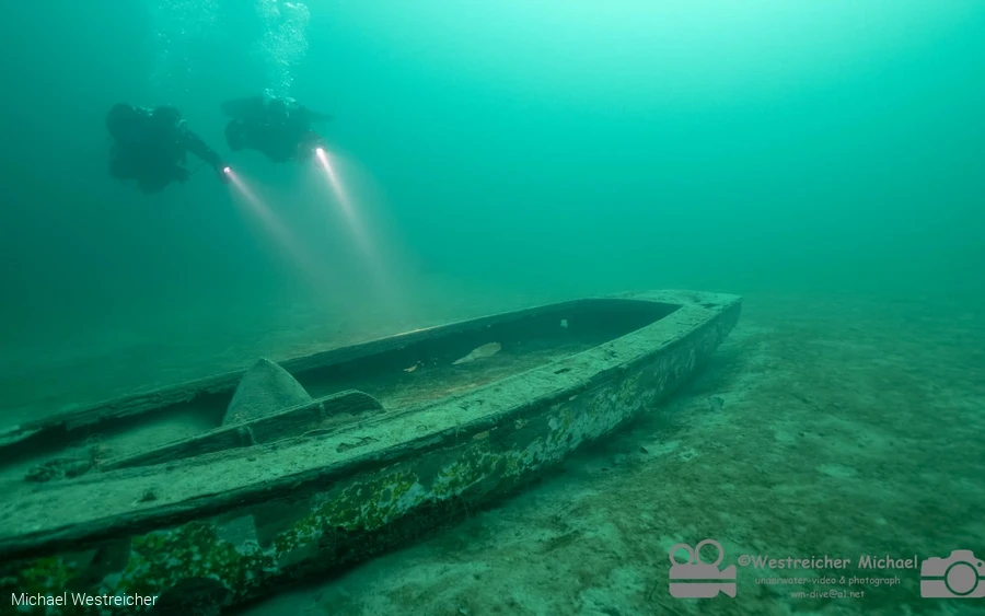 Tauchende Personen beleuchten ein versunkenes Boot auf dem Seegrund.