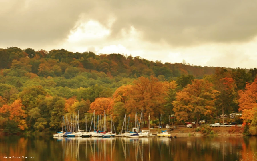 Segelboote am Ufer des Möhnesees im Herbst, Laubbäume in leuchtendem Rot und Orange spiegeln sich im Wasser.