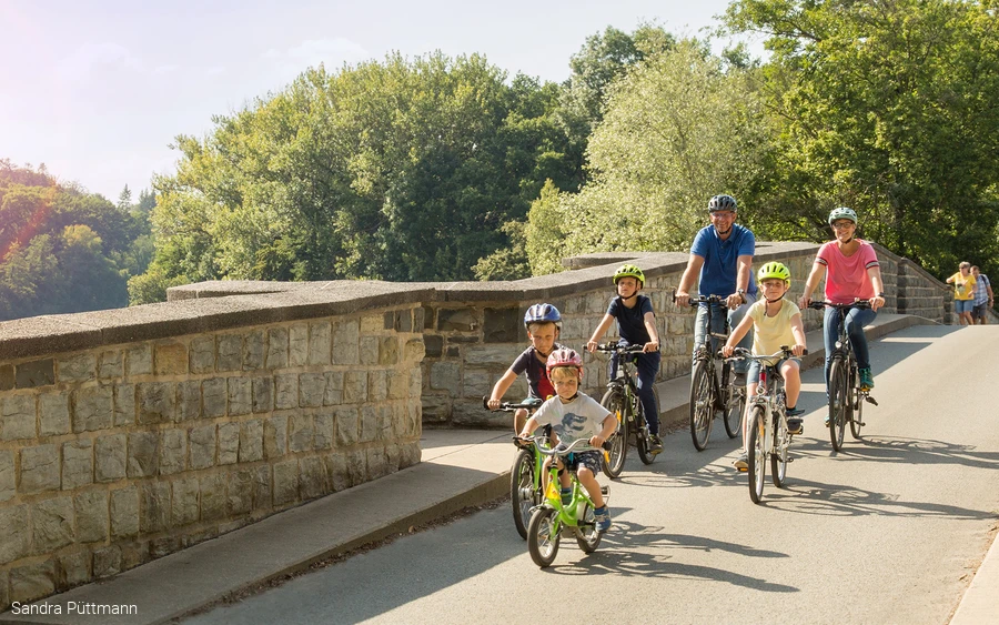 Familie mit Kindern fährt mit Fahrrädern über eine Steinbrücke am Möhnesee.