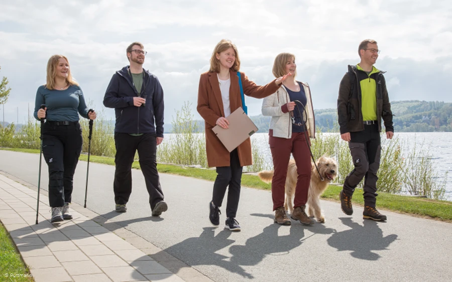 Gruppe von Personen spaziert mit Hund auf einem Weg am Seeufer.