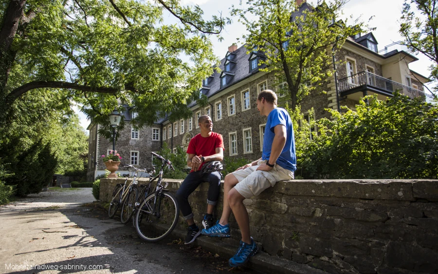 Zwei Männer mit Fahrrädern sitzen auf einer Steinmauer vor einem Haus mit Bäumen