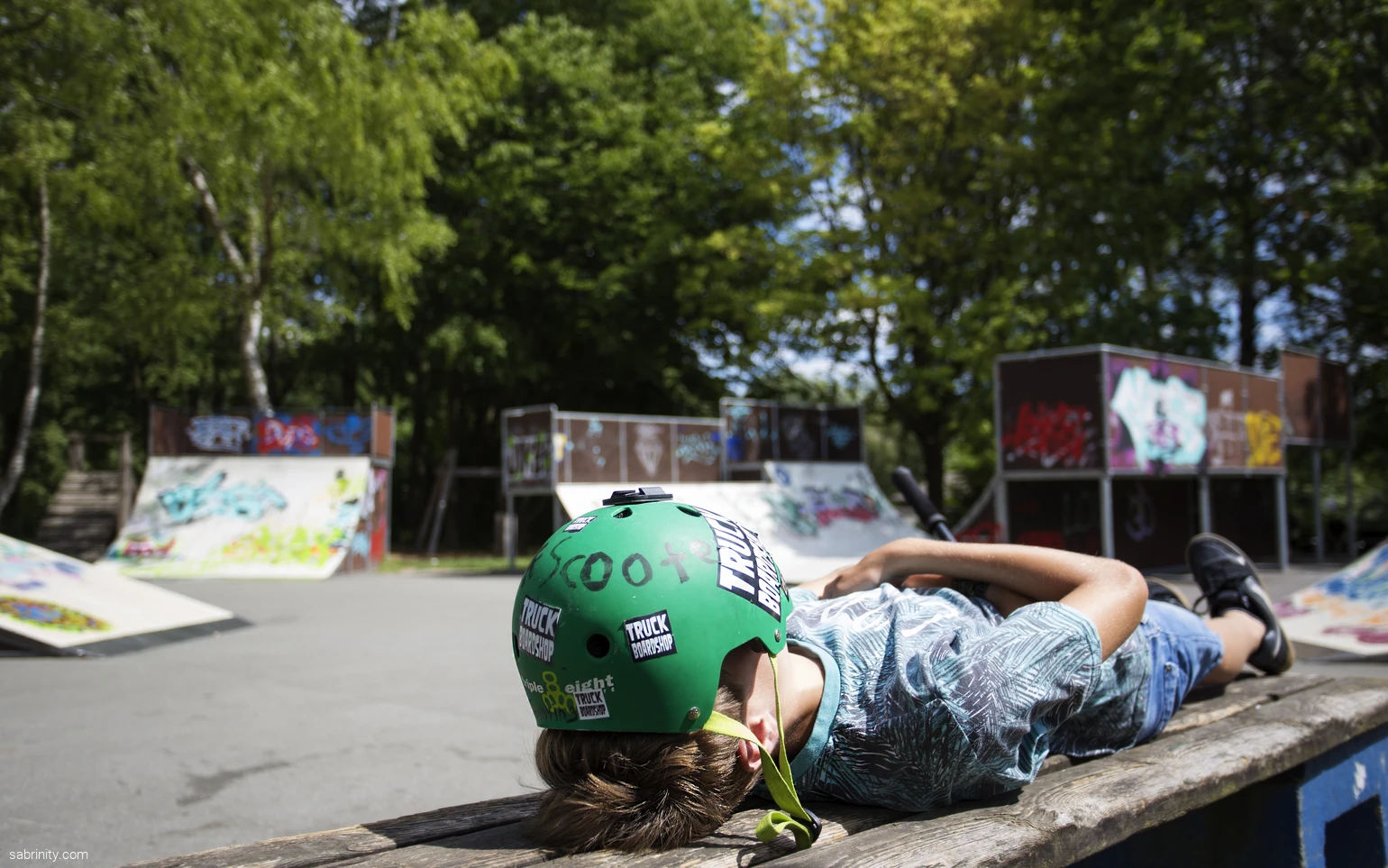 Junge Person mit grünem Helm liegt auf einer Bank in einem Skatepark.